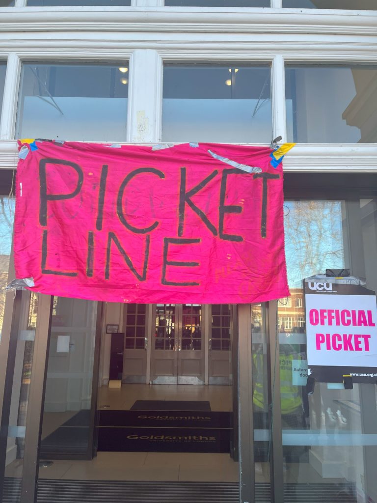 大学の本館に入る正面玄関前に作られたピケットライン。ガラスのスライドドアの上に"PICKET LINE"と書かれた濃いピンクの横断幕が掲げられ、横の扉には"OFFICIAL PICKET"と書かれた紙が貼られている。